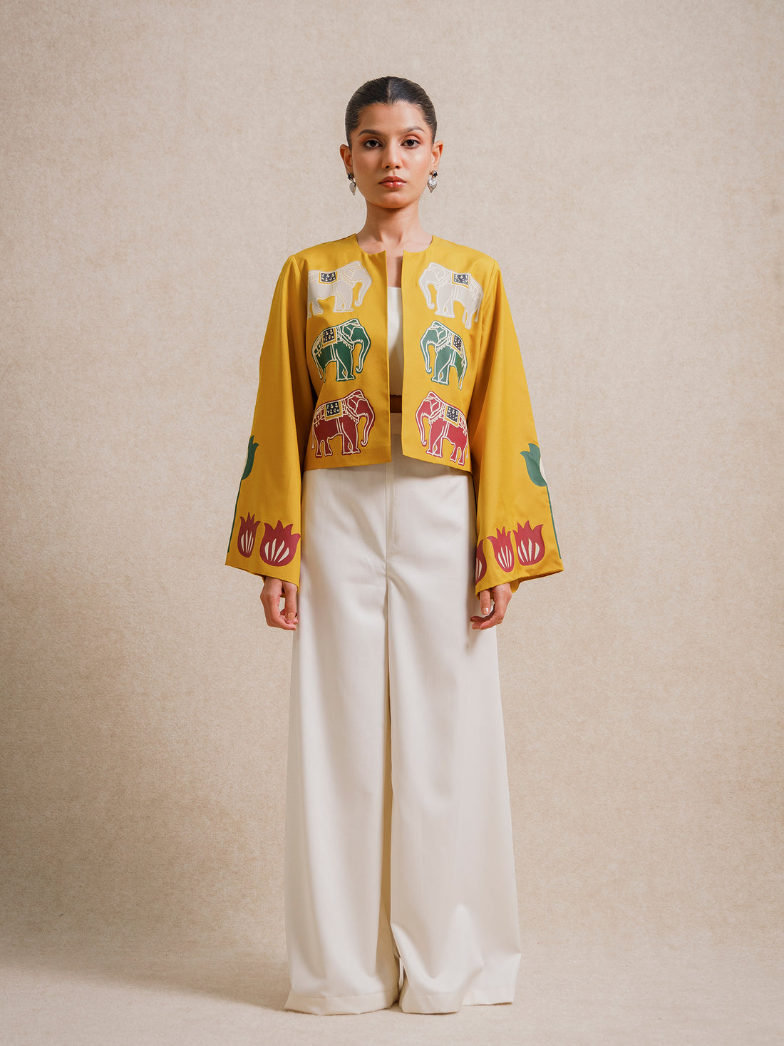 Front view of a model wearing the Alam Guman Ochre short jacket with hand-printed elephant and floral motifs, styled over a white outfit on a beige background