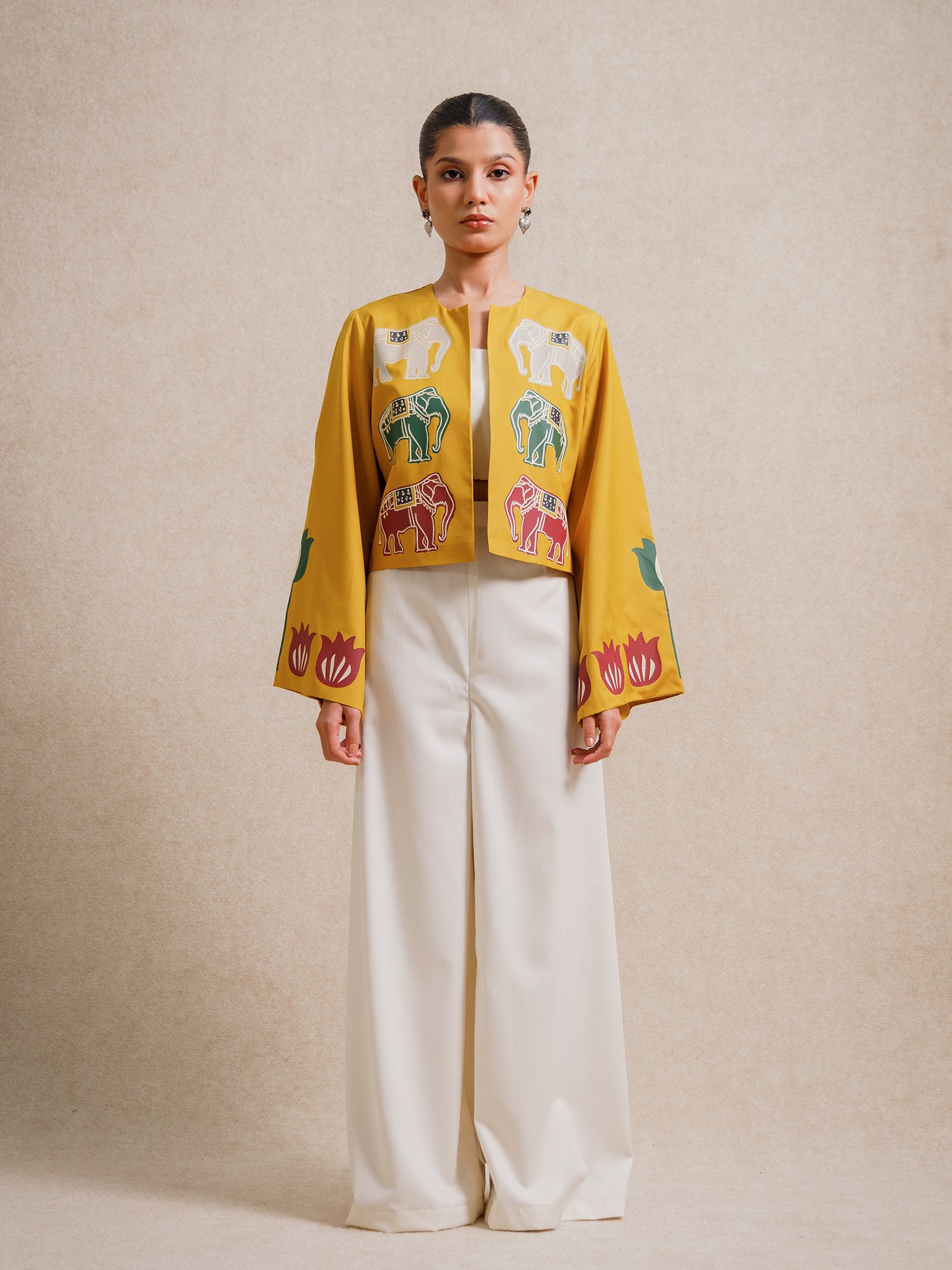 Front view of a model wearing the Alam Guman Ochre short jacket with hand-printed elephant and floral motifs, styled over a white outfit on a beige background