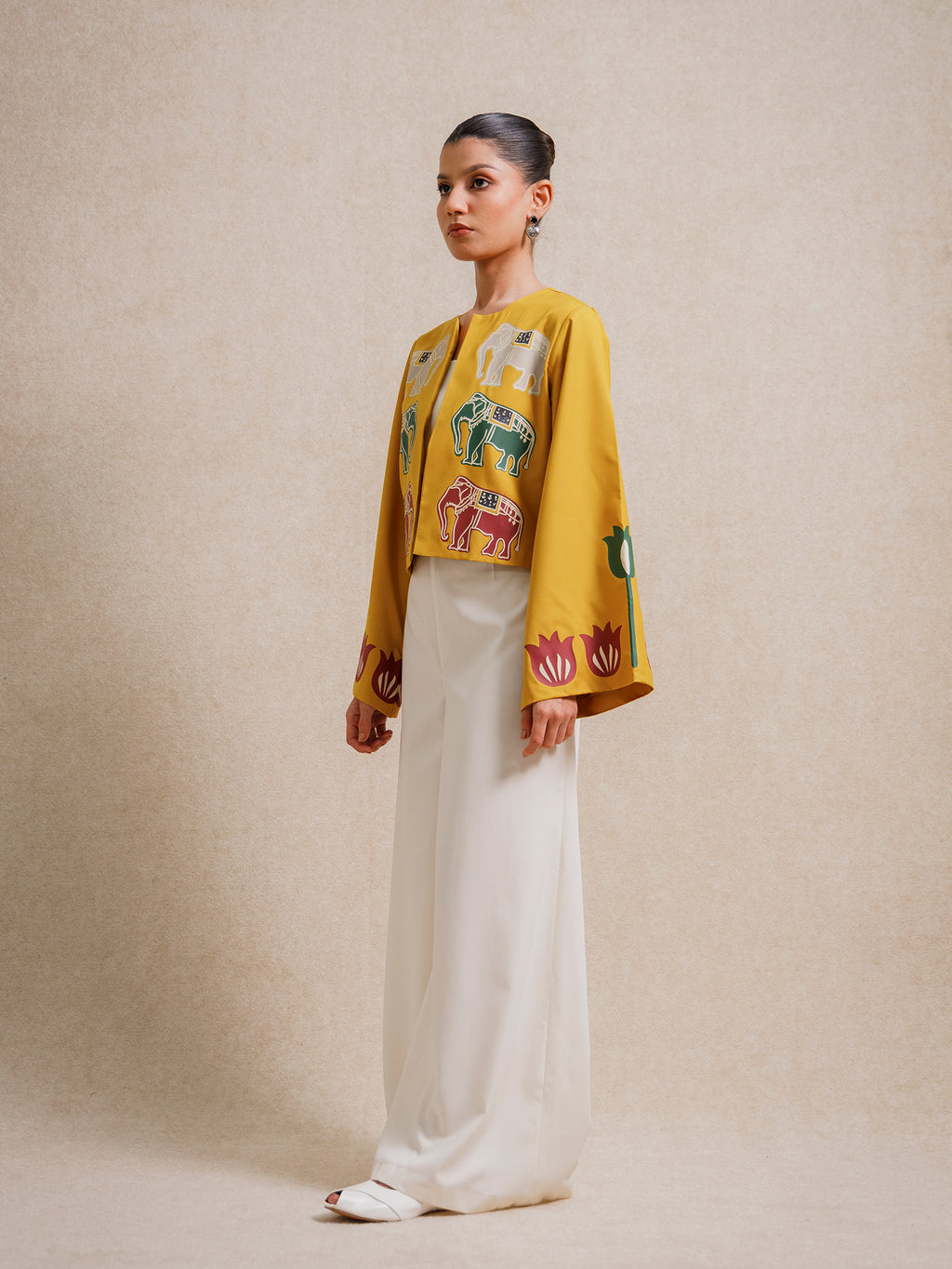 Side view of a model wearing the Alam Guman Ochre short jacket with hand-printed animal motifs, styled over a white outfit on a beige background