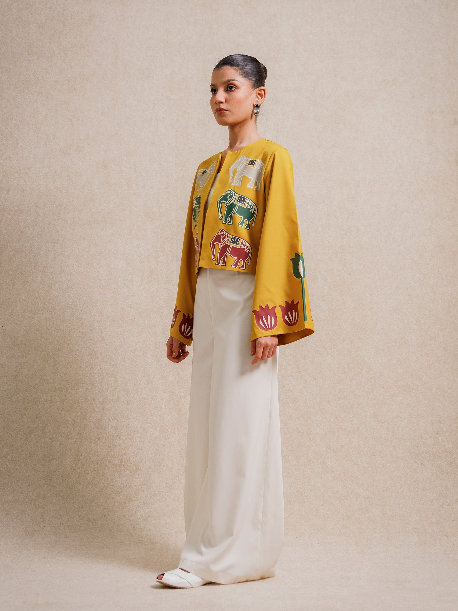 Side view of a model wearing the Alam Guman Ochre short jacket with hand-printed animal motifs, styled over a white outfit on a beige background